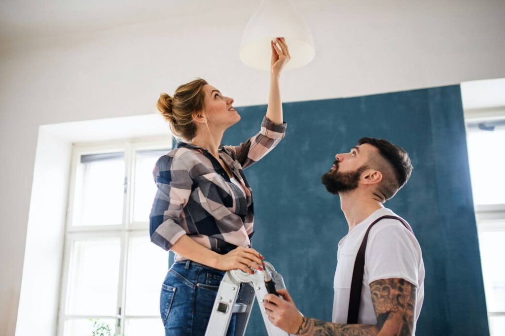 Two people changing a lightbulb on a ladder indoors.