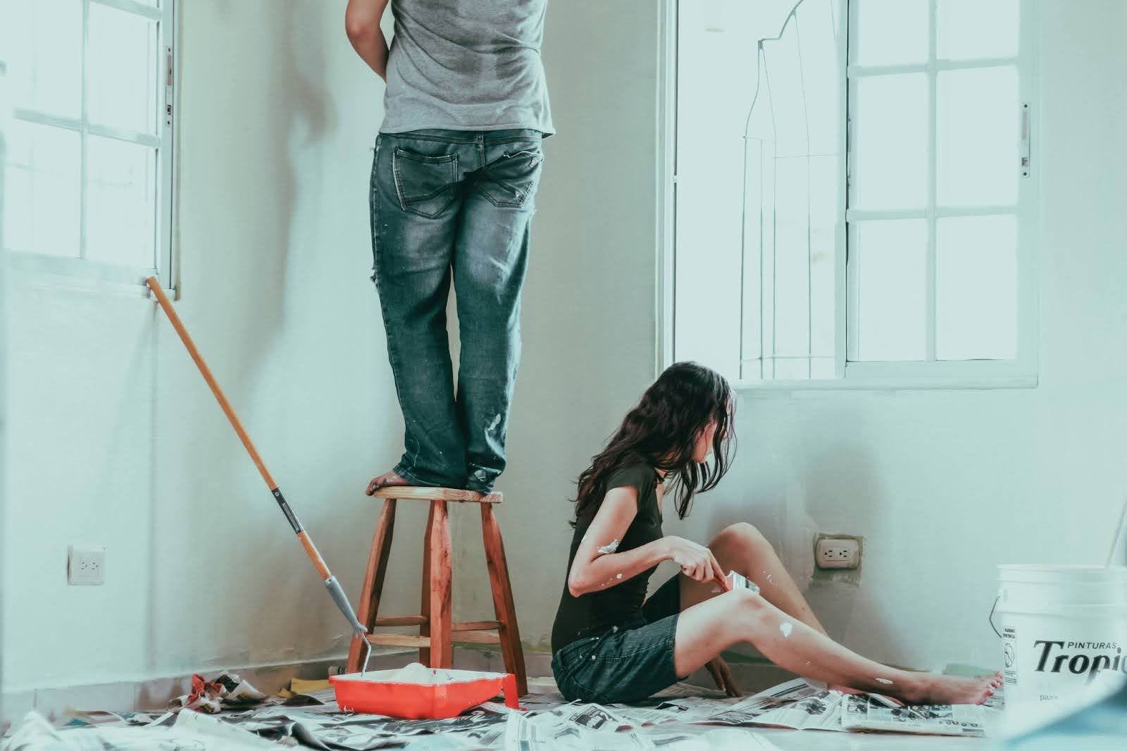 Two people painting a room with white walls and windows.