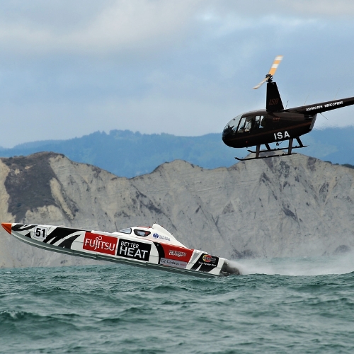 Speedboat racing on water with helicopter flying above, near rocky cliffs.